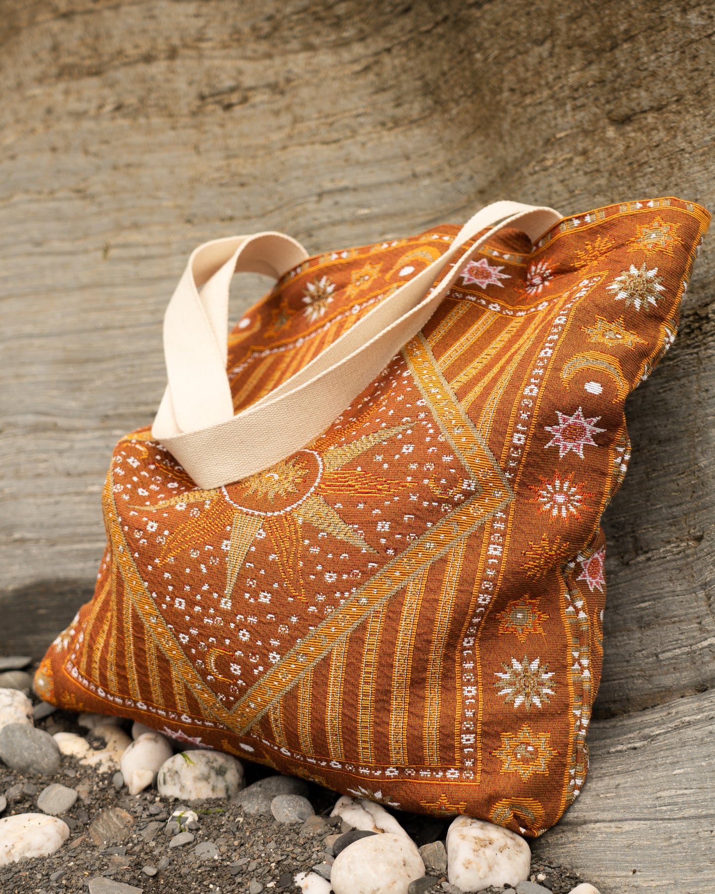 An orange and brown patterned fabric tote bag with cream canvas handles, resting on a stone beach with small white stones at the base.