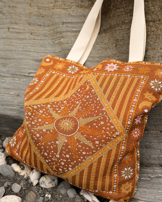 An orange and brown jacquard woven tote bag, featuring cream canvas shoulder straps, resting on beach with small stones at the base.