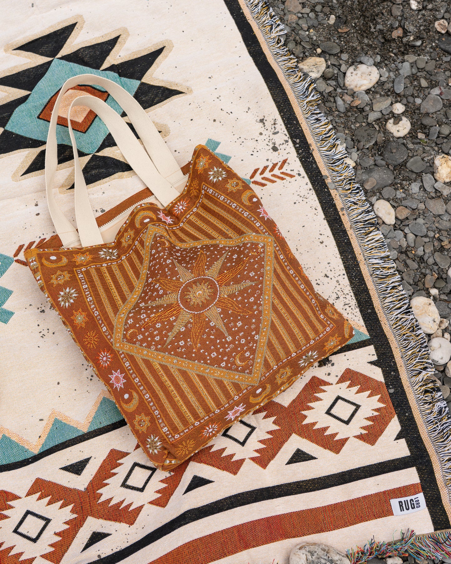 A handmade bohemian-style tote bag made from rust-orange and brown patterned fabric with cream canvas straps, laid flat on a colorful geometric rug next to a gravel surface.