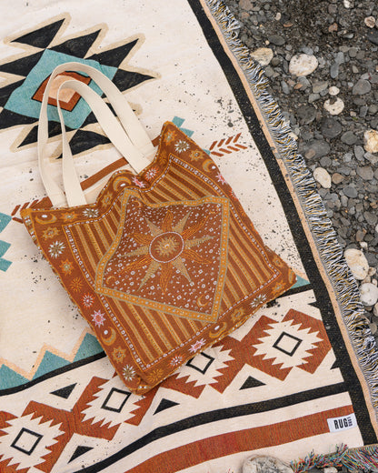A handmade bohemian-style tote bag made from rust-orange and brown patterned fabric with cream canvas straps, laid flat on a colorful geometric rug next to a gravel surface.